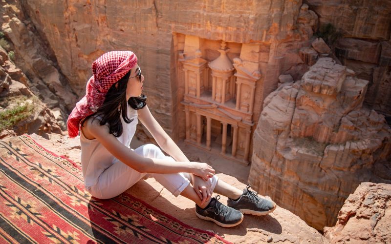 Asian woman traveler sitting on carpet viewpoint in Petra ancient city looking at the Treasury or Al-khazneh, famous travel destination of Jordan and one of seven wonders. UNESCO World Heritage site.