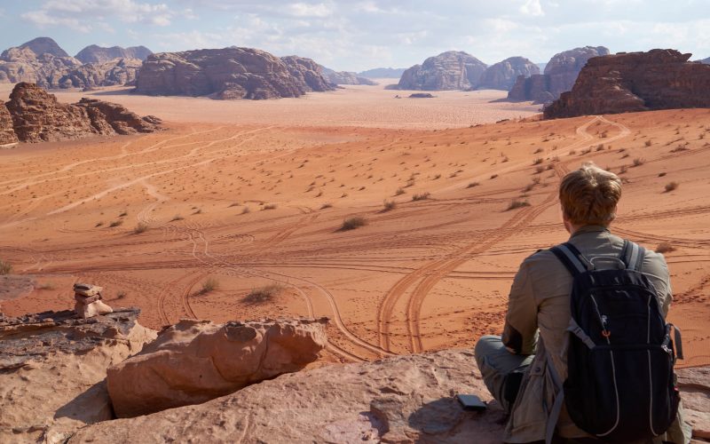 A traveler man sits on a cliff in the red desert of Wadi Rum in Jordan and admire the scenic landscape with dunes.
